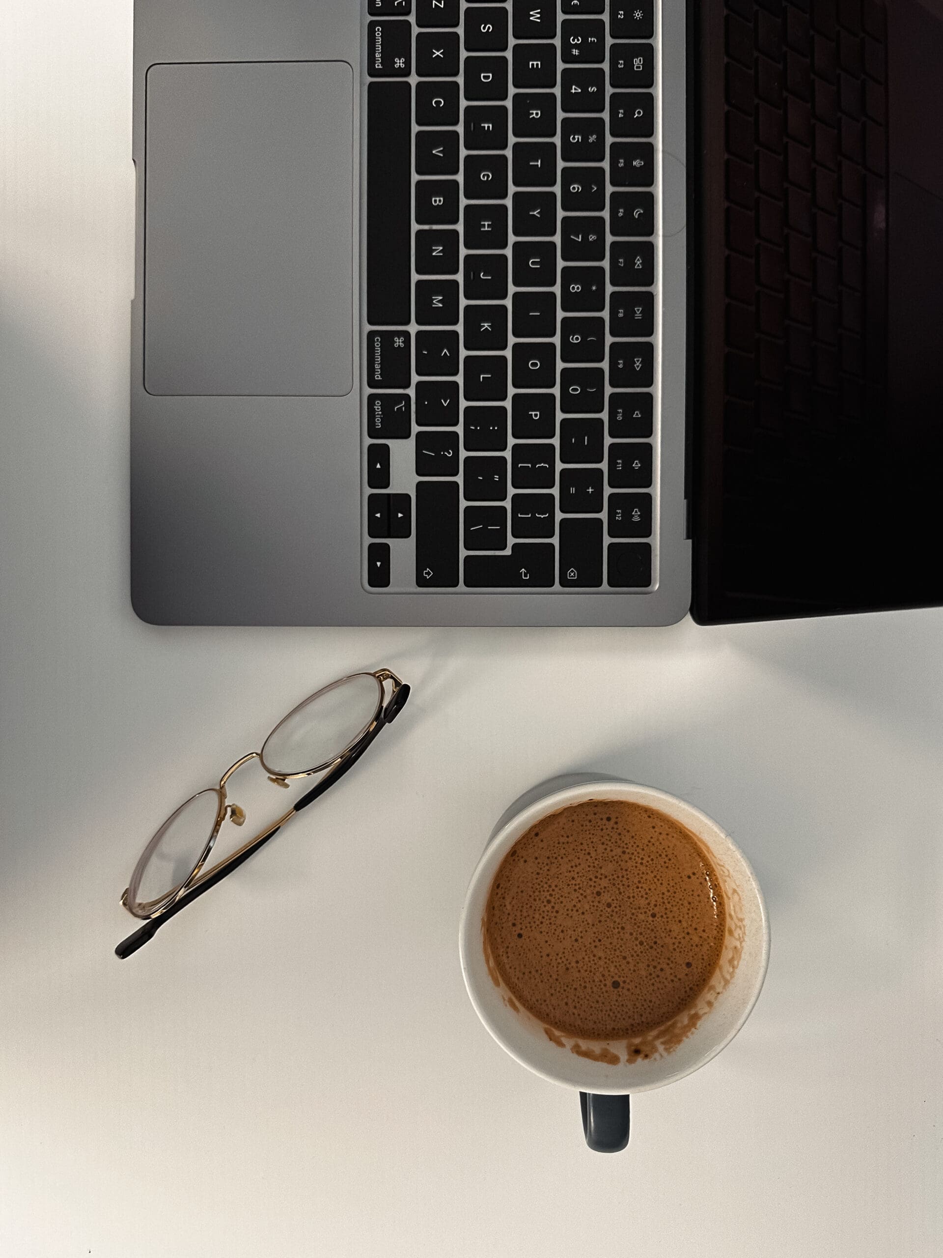 An overhead flatlay photo of a silver laptop, gold-rimmed glasses, and a mug of coffee with foam on a clean white desk, illustrating web design white space.