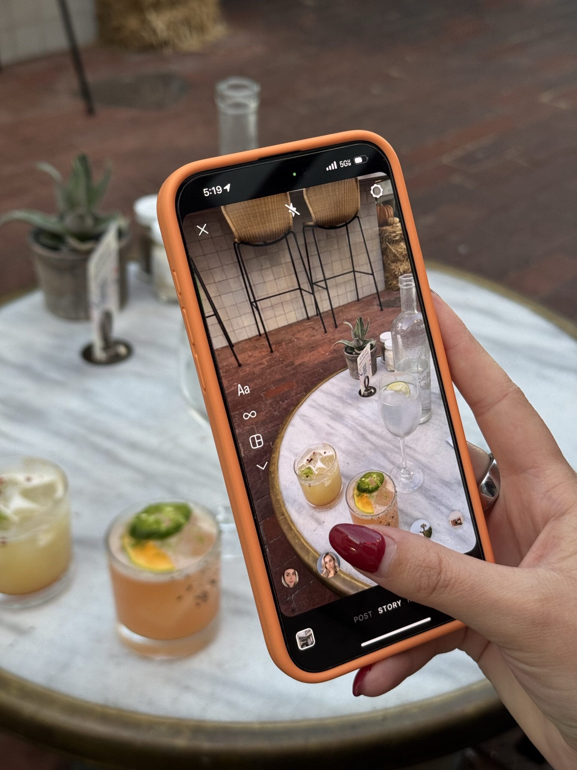 A person holding an orange-cased smartphone, capturing an Instagram Story photo of cocktails on an outdoor restaurant table.